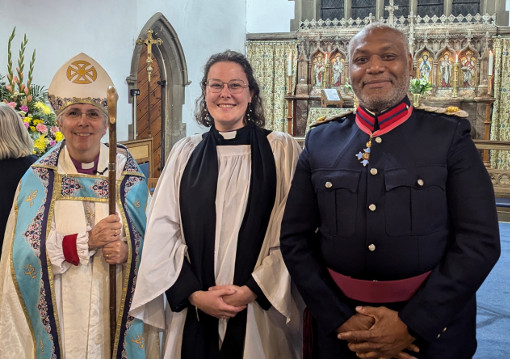 In front of St Paul's altar, Bishop Guli stands holding a crook and wearing an ornamented mitre with gold cross and a pale blue cloak embroidered with dark blue, red and gold, Revd Alexandra Podd in black cassock, white surplice and black tippet and Martin Griffiths in the uniform of the Deputy Lieutenant, Greater London, predominantly dark blue with red and blue collar and dark red belt and wearing a cross of pale blue stars at the neck.